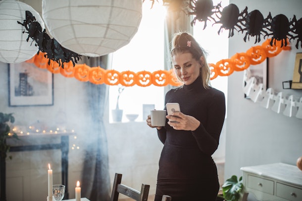 A woman in a black dress holds a cup of coffee and texts while standing in a living room that's decorated for Halloween.