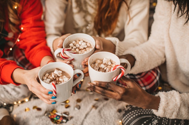 A group of friends holds mugs of hot cocoa, candy canes, and marshmallows together while sitting on a shaggy rug.