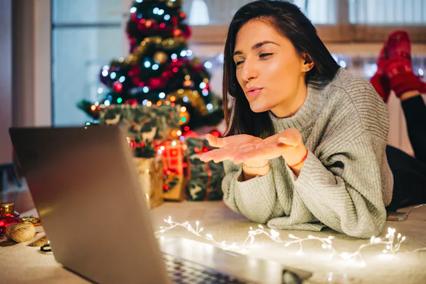 A girl lying down on fairy lights blows a kiss to her laptop screen during her virtual Christmas party.