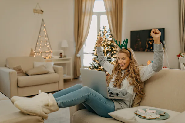 A woman on the couch, eating cookies and staying festive, raises her arms in celebration on her virtual Christmas party.