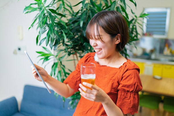 A young woman laughs while holding a drink and video chatting on her tablet. 