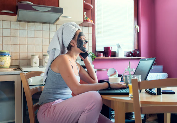 A young woman sits at her kitchen table and talks on the phone while doing a facial and wearing her hair in a towel.