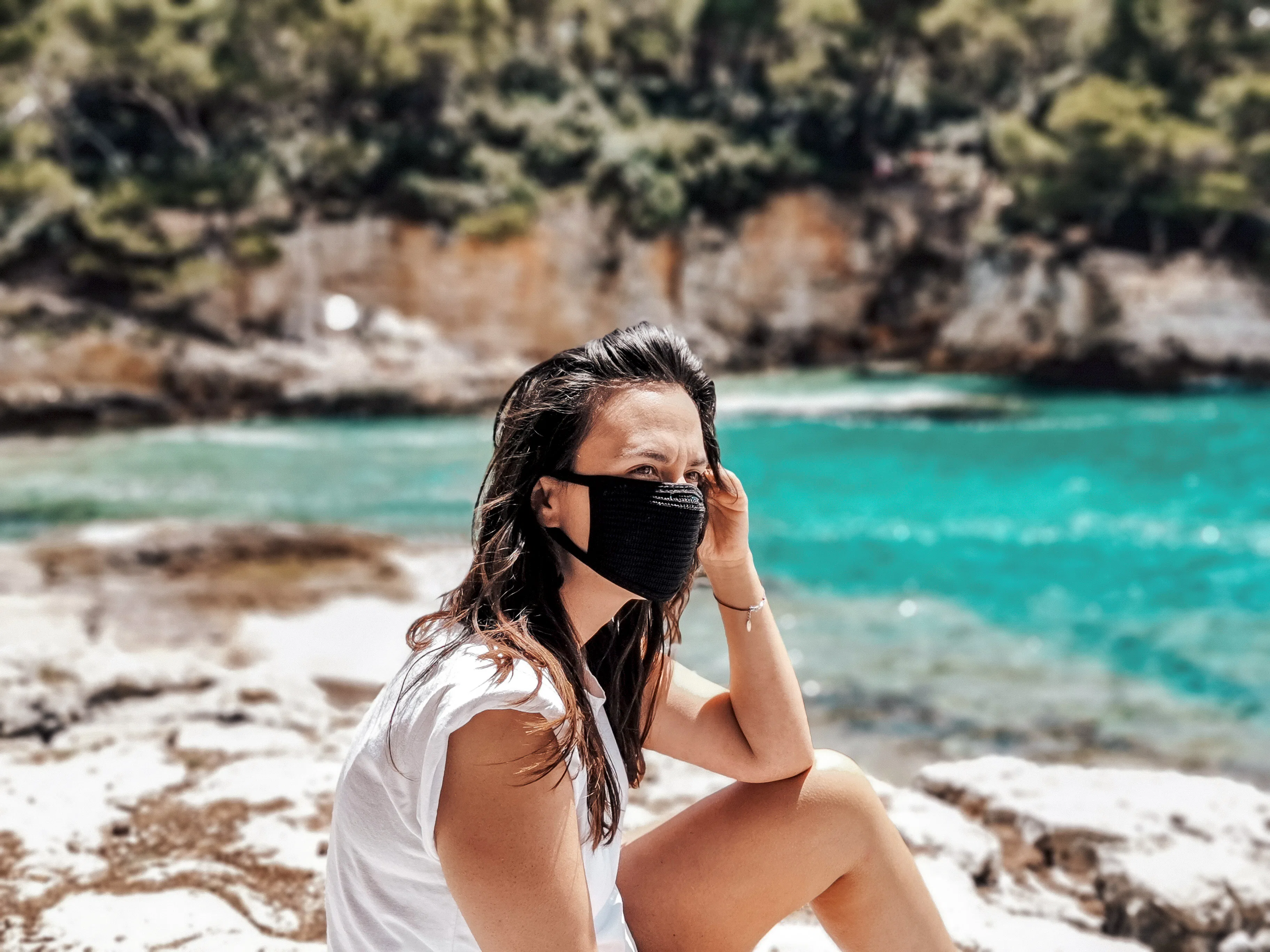 A young woman wears a black face mask while sitting next to a lake on a road trip.