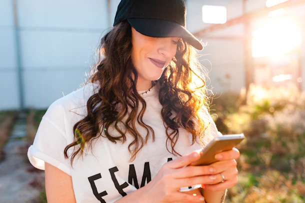 A happy woman wearing a baseball cap types on her phone.