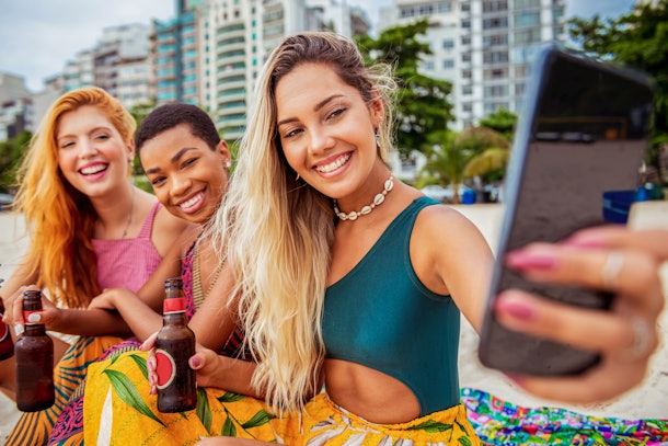 Three young women pose on the beach with their root beers and take a selfie.