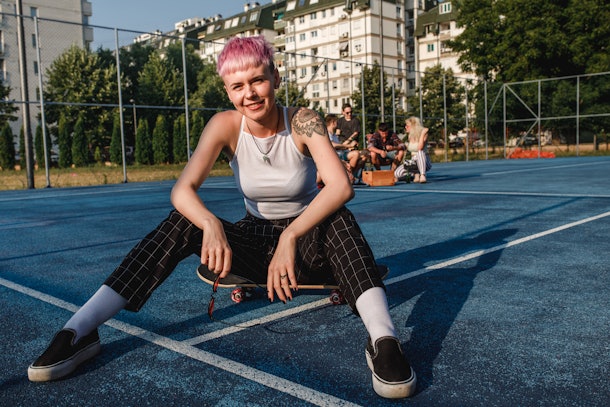 A young woman with short pink hair sits on a skateboard and poses for the camera at golden hour.
