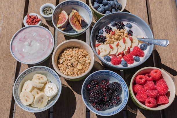 A smoothie bowl sits on a picnic table along with ingredients like fresh fruit, seeds, and granola to go into it.