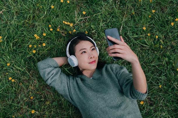 A young Asian woman looks at her phone and wears headphones while laying in the grass.