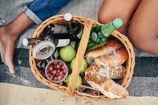Two women sit next to a picnic basket filled with bread, fruit, and drinks.