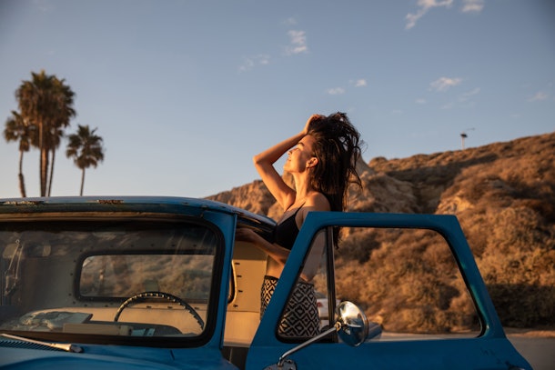 A young Asian woman runs her fingers through her hair while posing with a vintage car at sunset at the beach.