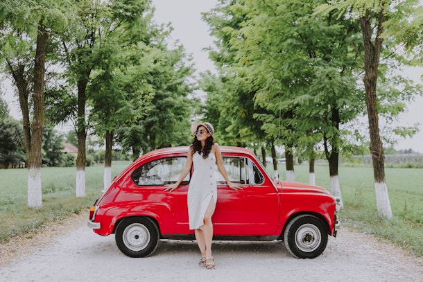 A young woman in a white dress poses against a red, classic car that's parked on a dirt road.