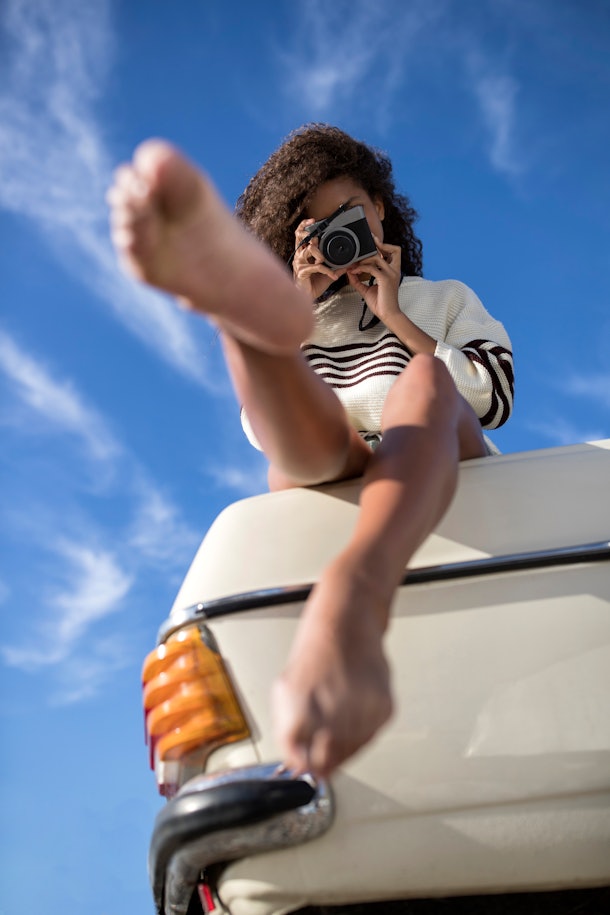 A young Black woman sits on the trunk of a vintage car and takes a photo on a vintage camera.