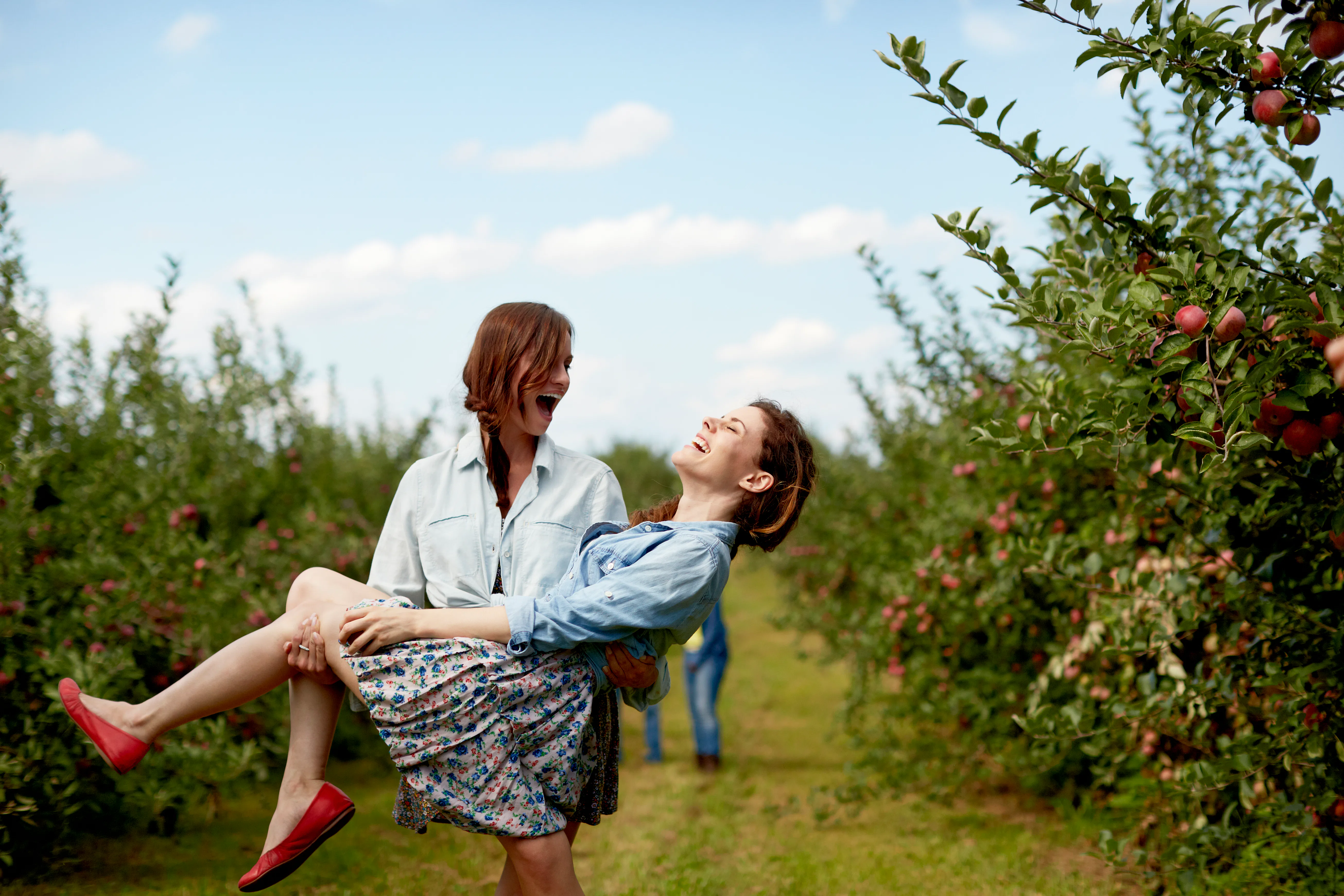 Two sisters dressed in button-down shirts and skirts laugh while at an apple orchard.