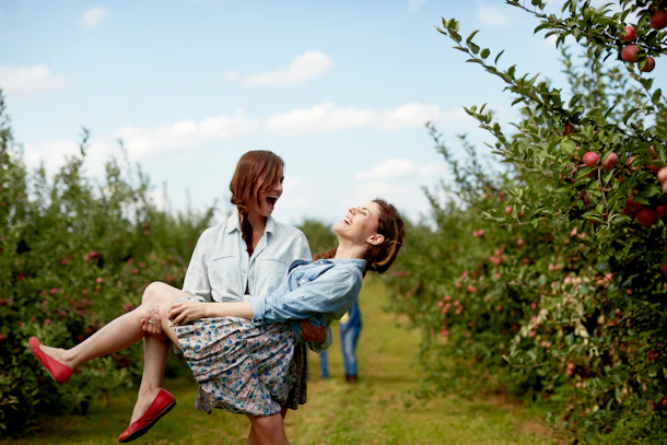 Two sisters dressed in button-down shirts and skirts laugh while at an apple orchard.