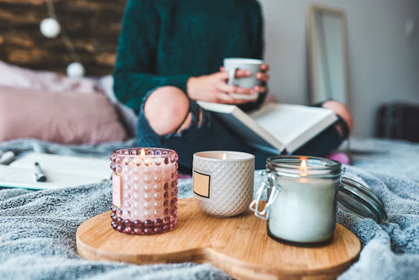 A young woman sits next to three candles in bed while reading a book and drinking tea.
