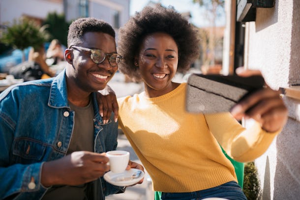 Happy smiling African American couple, sitting in a coffee shop, relaxing and enjoying their vacation and each other, taking a selfie together with a smart phone