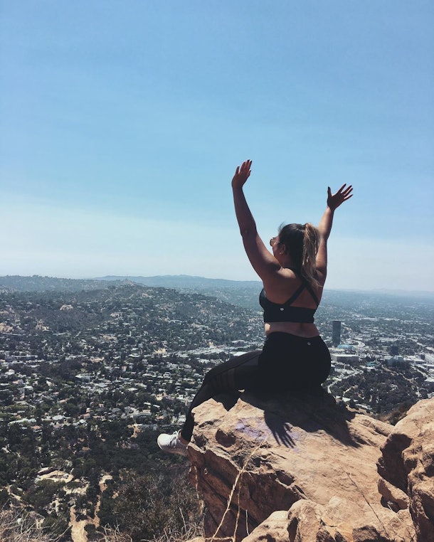 A girl with her back turned to the camera sits on a cliff with her arms raised overlooking a city below.
