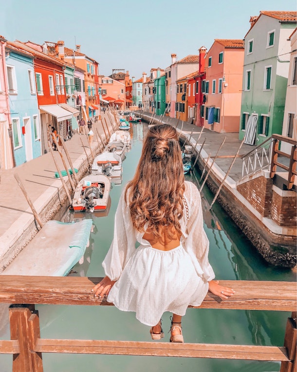 A brunette woman in a white sundress sits on a bridge overlooking a canal and colorful buildings in Italy.
