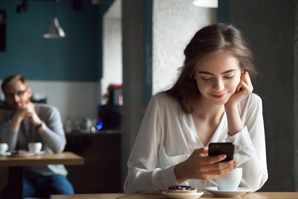 Young curious guy likes millennial beautiful girl looking with interest watching smiling lady sitting nearby in cafe using smartphone, flirt in public place, dating and love at first sight concept