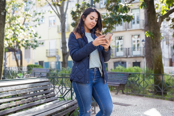 Serious woman using smartphone and standing outdoors. Pretty young lady wearing jacket with trees and buildings in background. Urban lifestyle and communication concept.