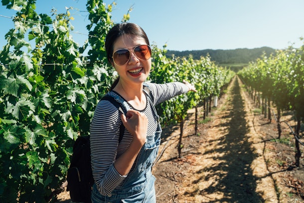asian woman standing in napa valley vineyard cheerfully showing camera the blooming plants trees in spring. young girl in sunglasses pointing finger in grape winery sunny day. female visitor smiling