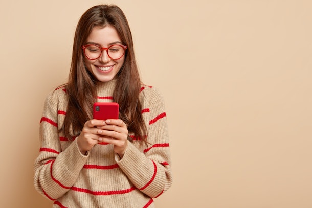 Indoor photo of satisfied teenage girl texts on cellular, reads interesting article online, wears casual outfit, creats new publication on own web page, isolated over brown studio wall with free space