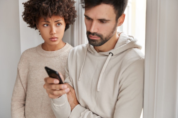 Attractive man wears hoodie, ignores live communication with wife, focused in modern smart phone, reads news online, sad dark skinned woman feels jealous as her husabnd messages with someone