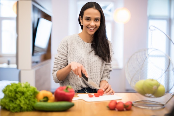 The young woman cuts vegetables in the kitchen with a knife and laptop on the table. Vegetable Salad. Diet. Dieting Concept. Healthy Lifestyle. Cooking At Home. Prepare Food. With place for text
