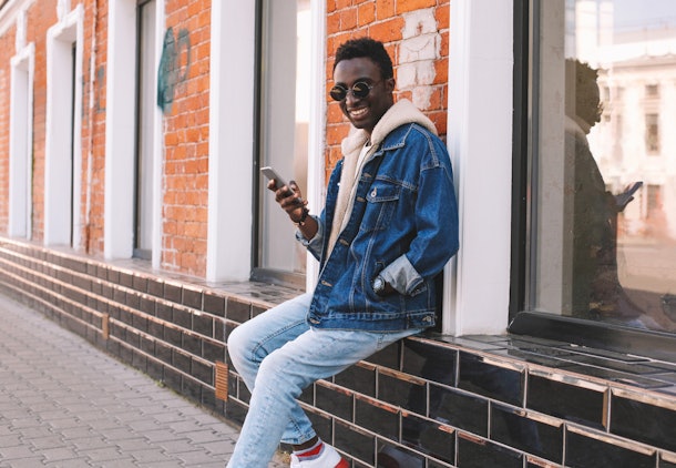 Happy cool smiling african man with smartphone sitting on city street over brick wall background