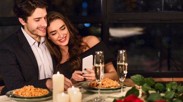 A happy brunette couple looks down at photos on a phone during a romantic dinner with champagne and spaghetti.