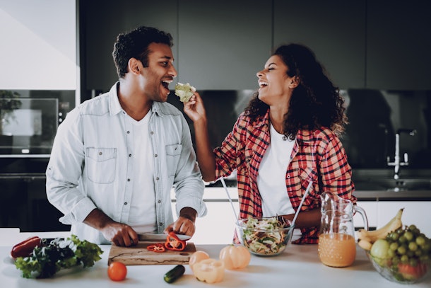 A happy couple cooking makes salad at a white kitchen counter, while the girl laughs and feeds her boyfriend food.