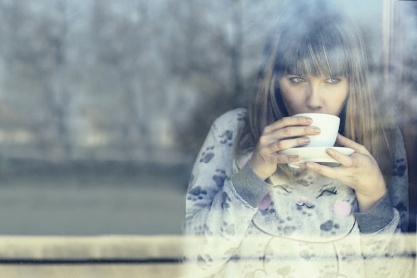 young woman in night dress drinking warm coffee or tea inside home looking out through the window glass on cold winter morning. concept of lazy winter morning and comfort of a cozy appartment.