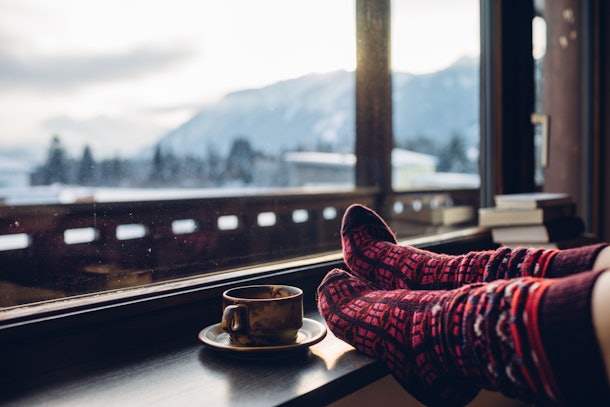 Feet in woollen socks by the Alps mountains view. Woman relaxes by mountain view with a cup of hot drink. Close up on feet. Winter and Christmas holidays concept.