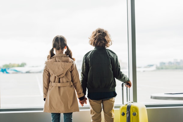 back view of kids holding hands, standing near window in waiting hall in airport