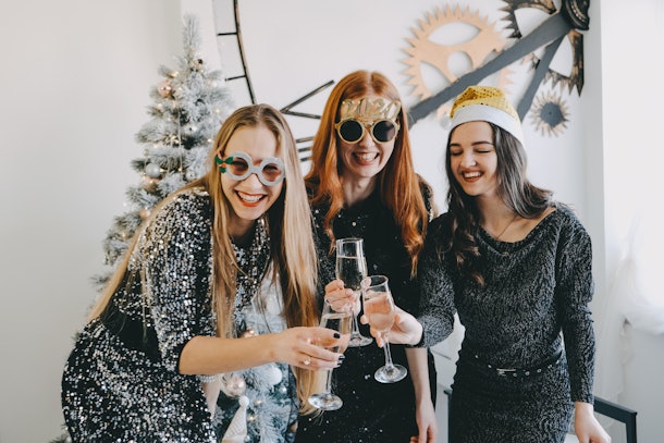 Cheers! New Year Eve, Christmas Celebration. Three partying girls clinking flutes with champagne. Young beautiful women in evening dress celebrating party, drinking wine and dancing.