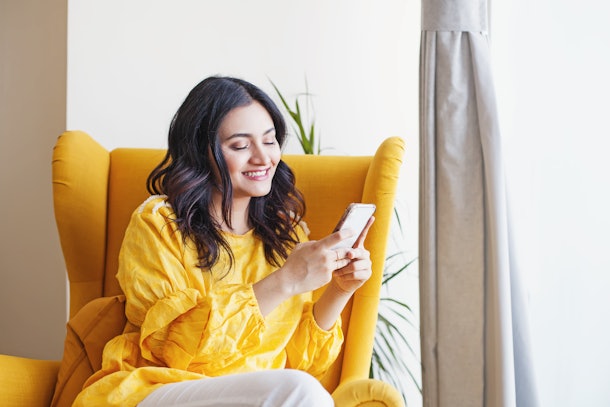 Young beautiful Indian woman using her mobile phone while sitting in the armchair at home