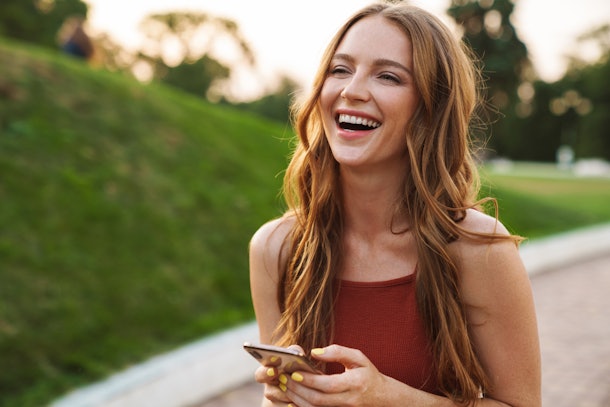 Photo of a happy laughing emotional young ginger woman walking in nature green park outdoors using mobile phone.