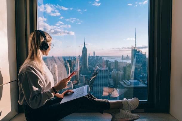 Blonde hair female blogger listening to music in headphones during freelance work on laptop computer. Hipster girl traveler enjoying panoramic view of New York downtown and studying with audio book
