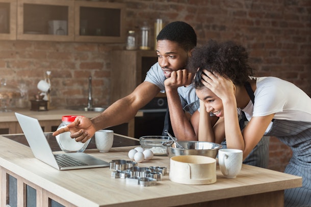 Shocked african-american couple baking with recipe on laptop in loft kitchen