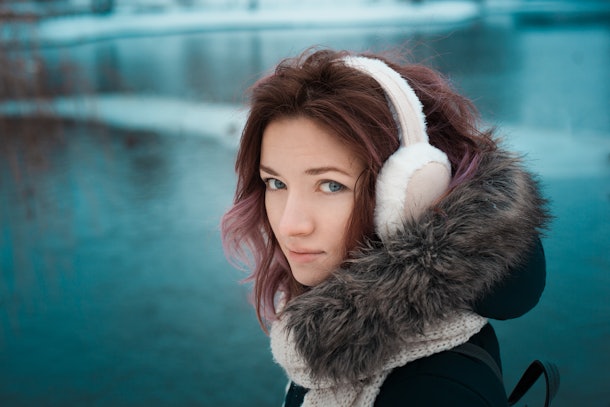 girl with red hair in a dark winter jacket and hood with fur in fur pink headphones in the park against the backdrop of the lake
