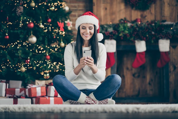 En casa, la chica positiva con decoraciones X-Mass usa un sombrero de Papá Noel con descuento de Navidad en busca de sus mensajes de texto tipo teléfono inteligente en la cómoda alfombra del piso.