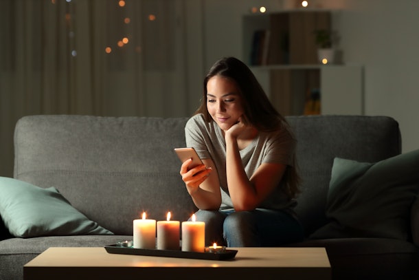 Relaxed girl using phone in the night with candle lights sitting on a couch in the living room at home