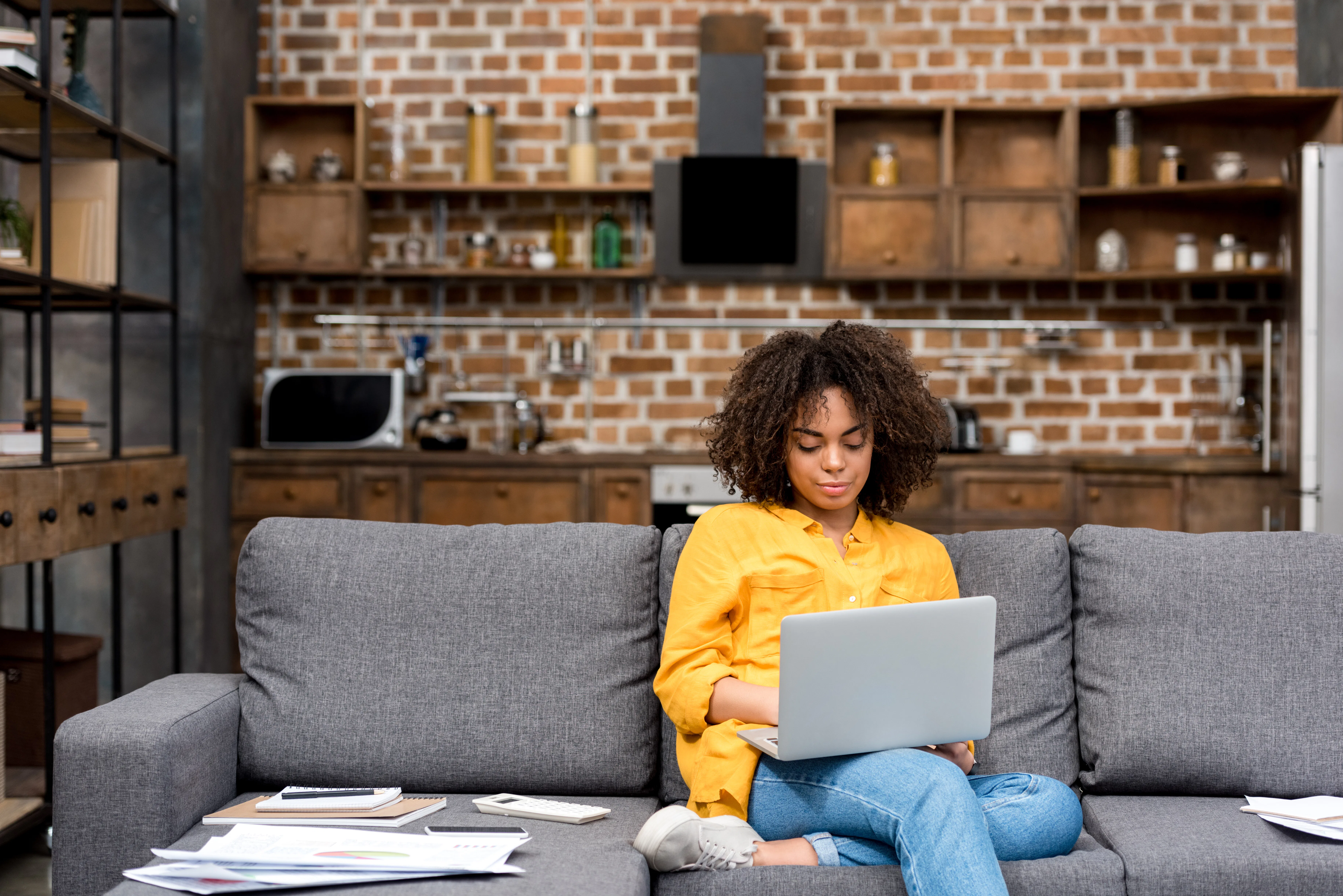 A woman in a yellow button-down sits on her couch with her laptop and papers nearby. 