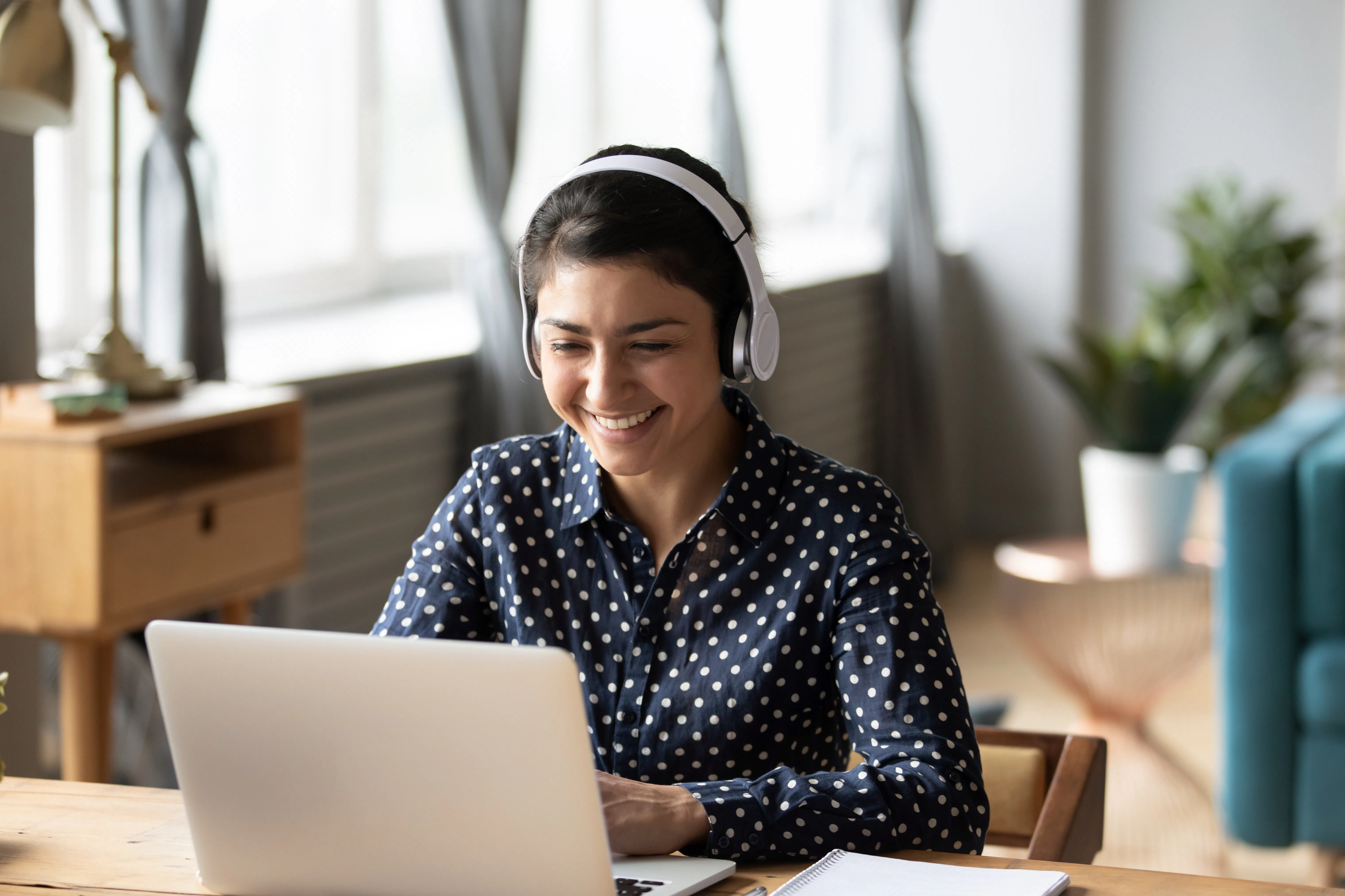 A happy woman wearing headphones and a blue polka dot button down shirt smiles while typing on her laptop.