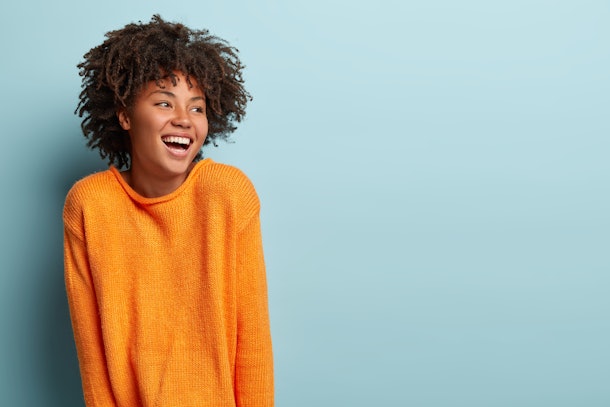 Glad overemotive dark skinned lady with curly hairstyle, laughs happily, expresses sincere emotions, being amused by friend, dressed in orange casual jumper, models in studio alone with mockup space