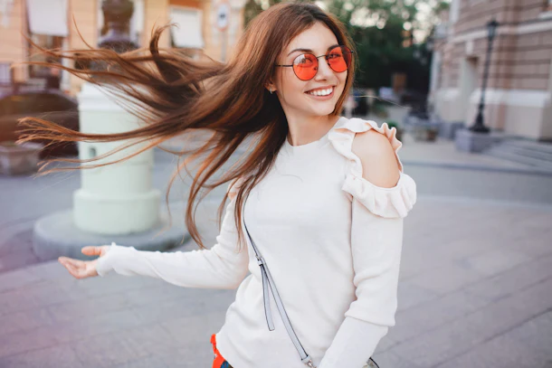 A happy brunette woman flips her hair, dressed in red sunglasses and a white sweater.