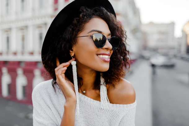 A fashionable woman wearing an off-the-shoulder white sweater, dangly white earrings, sunglasses, and a black felt hat smiles and looks off into the distance.