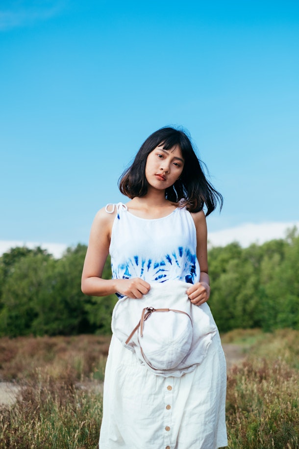 A young Asian woman wears a tie-dye tank top while standing for a photo in a grass field on a sunny day.